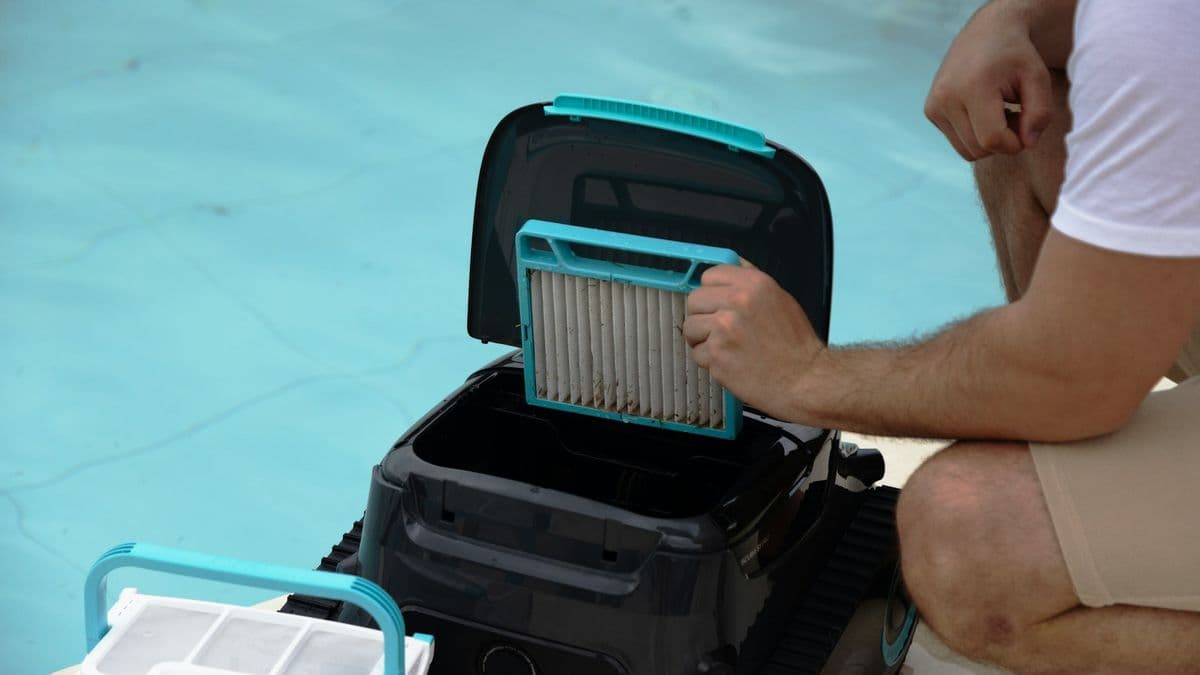 A technician pulling a debris-filled filter cartridge from a robotic pool cleaner beside the water